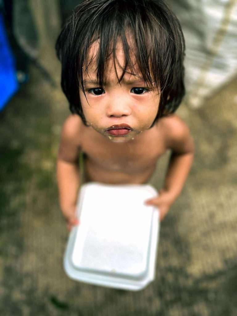 Child holding a white container, looking solemn, in a poverty-stricken area of Tondo, reflecting the need for educational support and feeding programs.