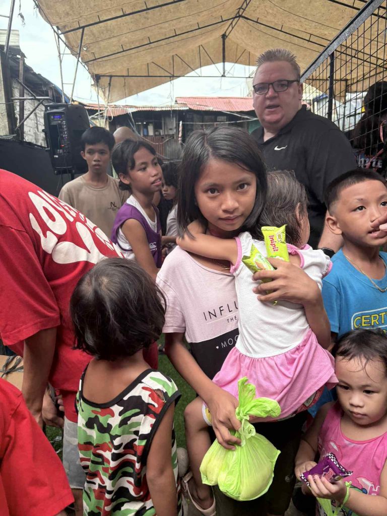 Children in Tondo, Manila, with a girl holding a younger child, surrounded by peers, receiving food and support from local volunteers.