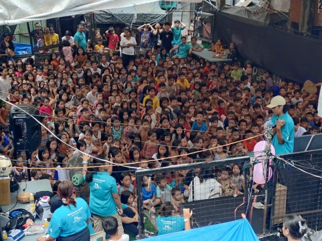 Crowd of children and adults gathered at a community event in Tondo, Manila, with volunteers in blue shirts engaging the audience, highlighting local outreach efforts for education and support.