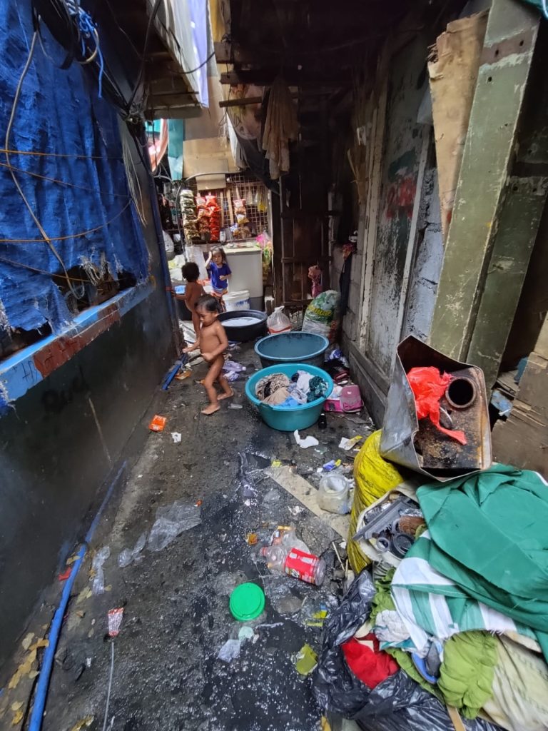 Children playing in a narrow, littered alley in Tondo, Manila, surrounded by trash and makeshift living conditions, highlighting the poverty and struggles faced by families in the community.